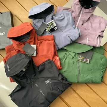 A pile of shell jackets in orange, green, lilac and grey on a wooden floor.